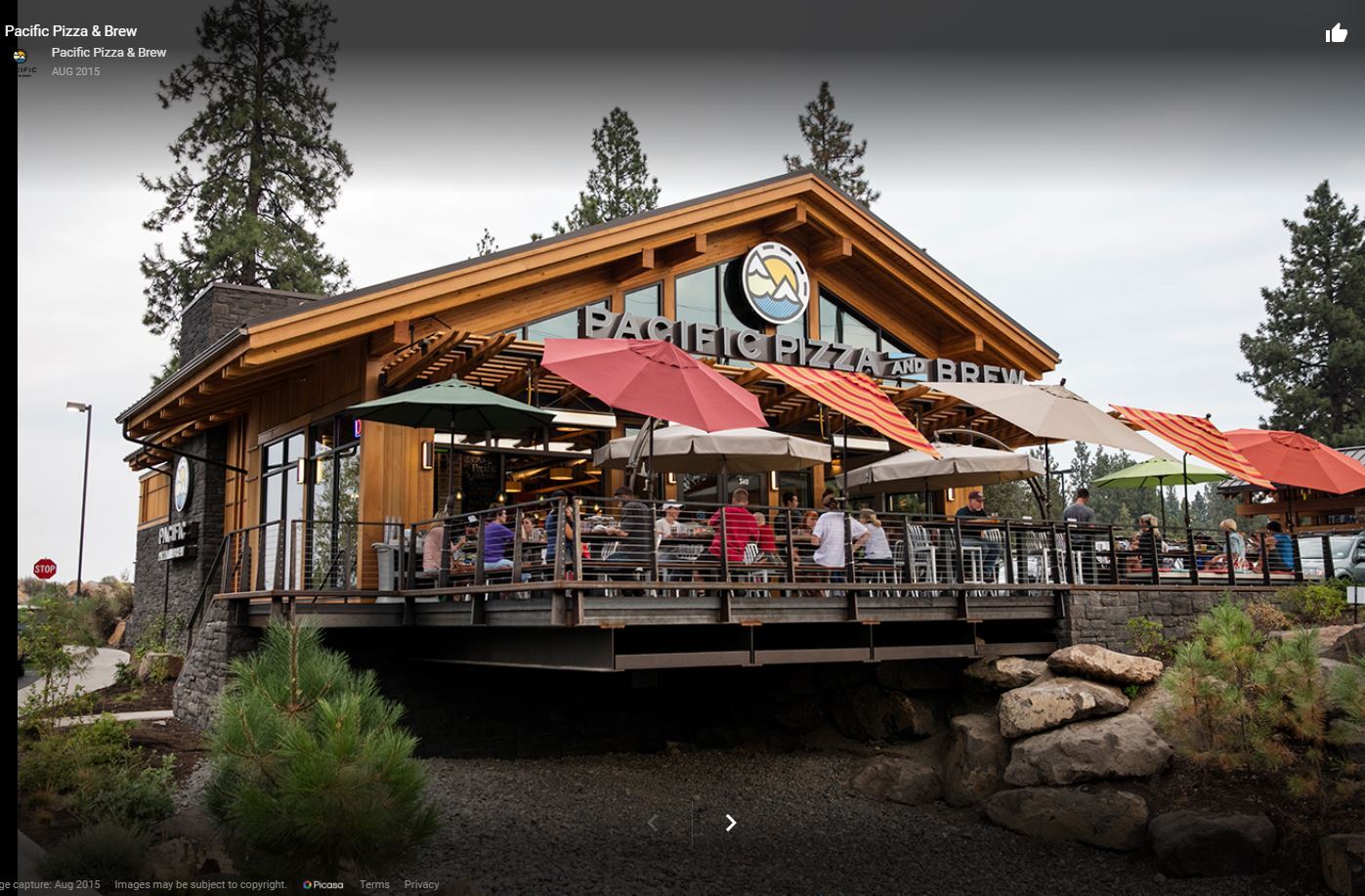 Outdoor dining area of Pacific Pizza & Brew, featuring patrons under colorful umbrellas on a wooden deck, surrounded by trees.