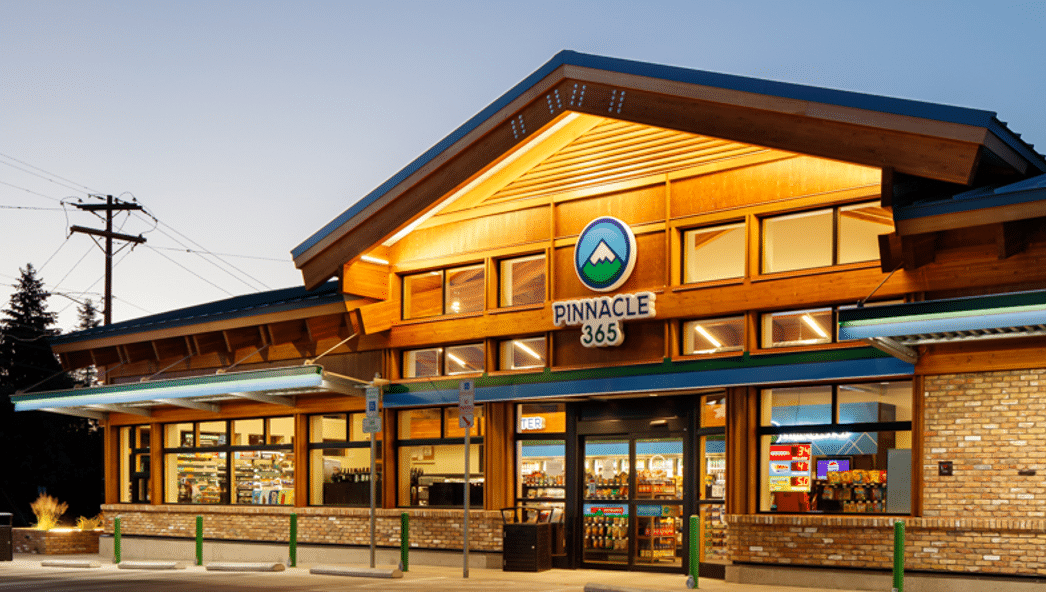 Pinnacle 365 store exterior at dusk, showcasing its wooden architecture, illuminated signage, and stocked shelves visible through large windows.