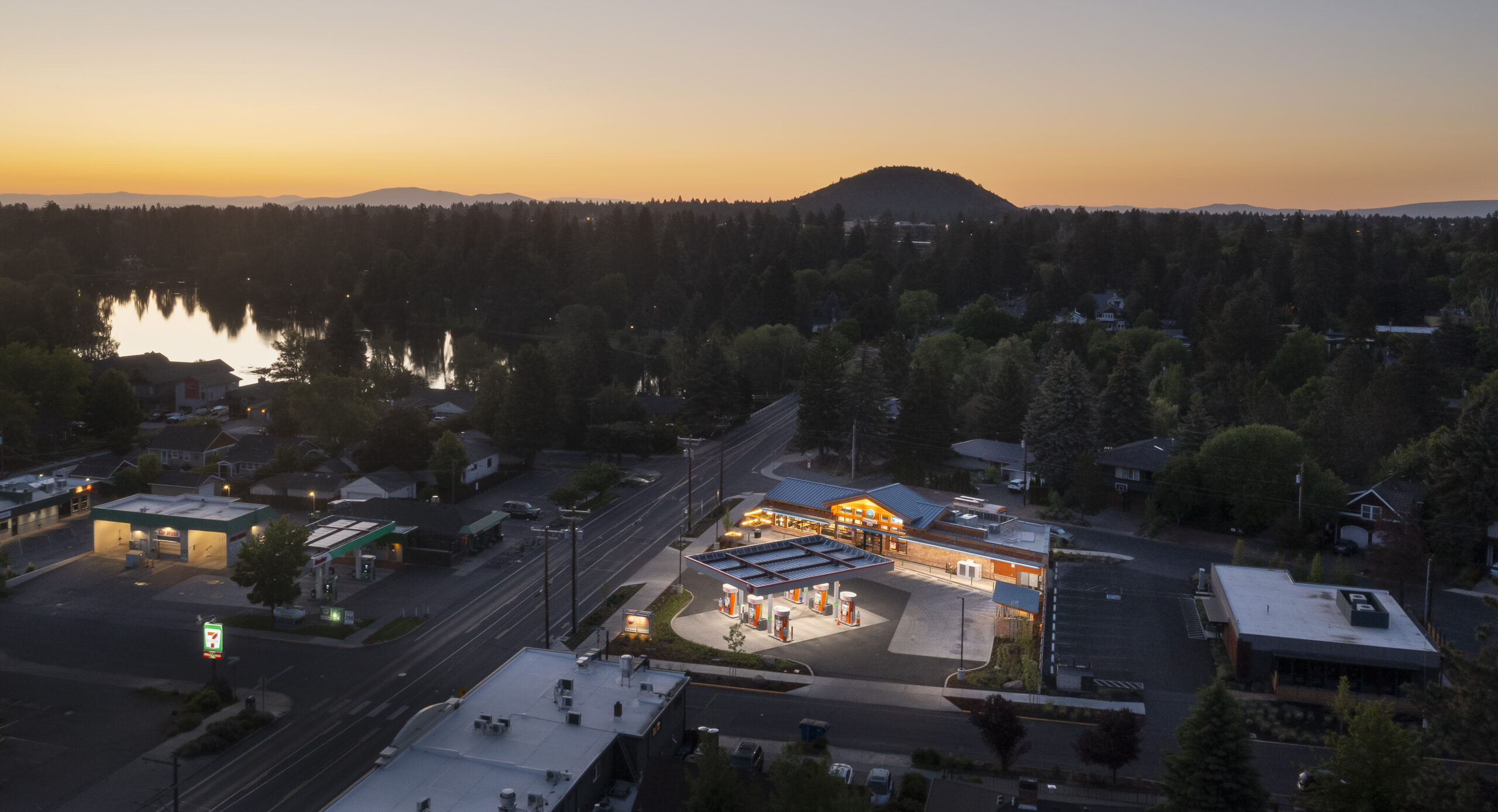 Aerial view of a gas station and convenience store illuminated at dusk, surrounded by trees and residential areas, with a lake reflecting the sunset.