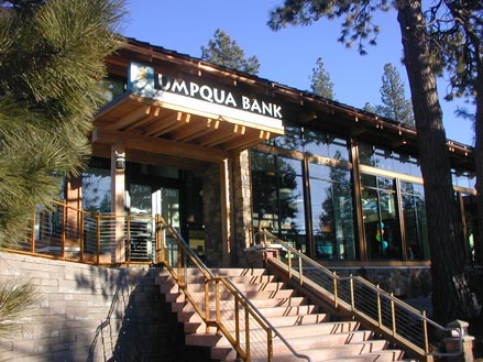 Modern Umpqua Bank building with large windows and wooden accents, surrounded by trees. Stairs lead to the entrance, highlighting its welcoming design.