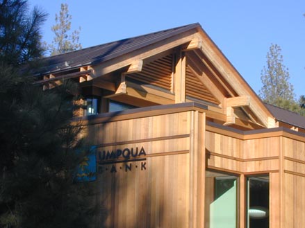 Umpqua Bank building featuring a modern wooden design with large windows and a distinctive roofline, set against a clear blue sky and trees.