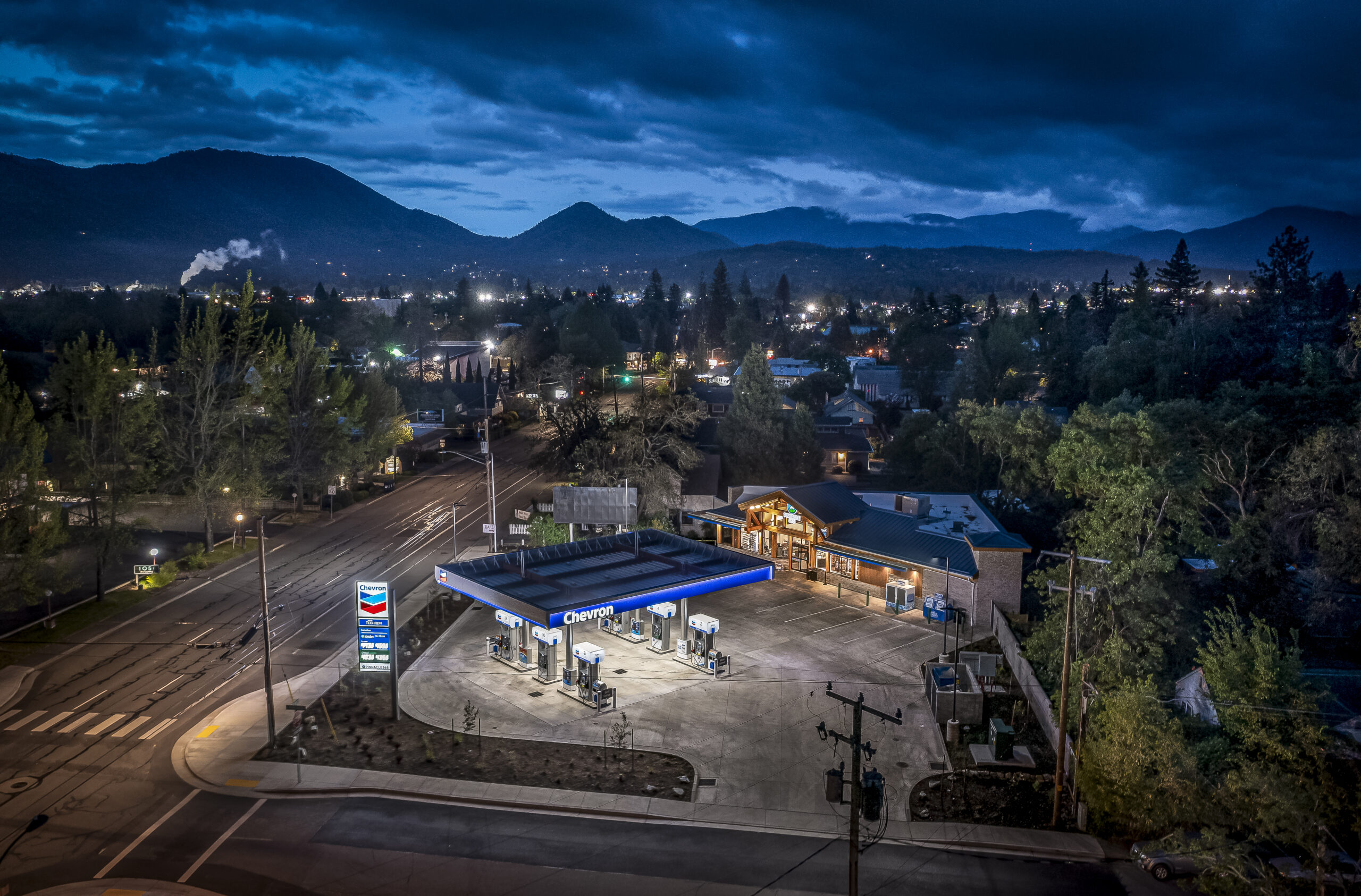 Chevron gas station illuminated at night, surrounded by mountains and trees.