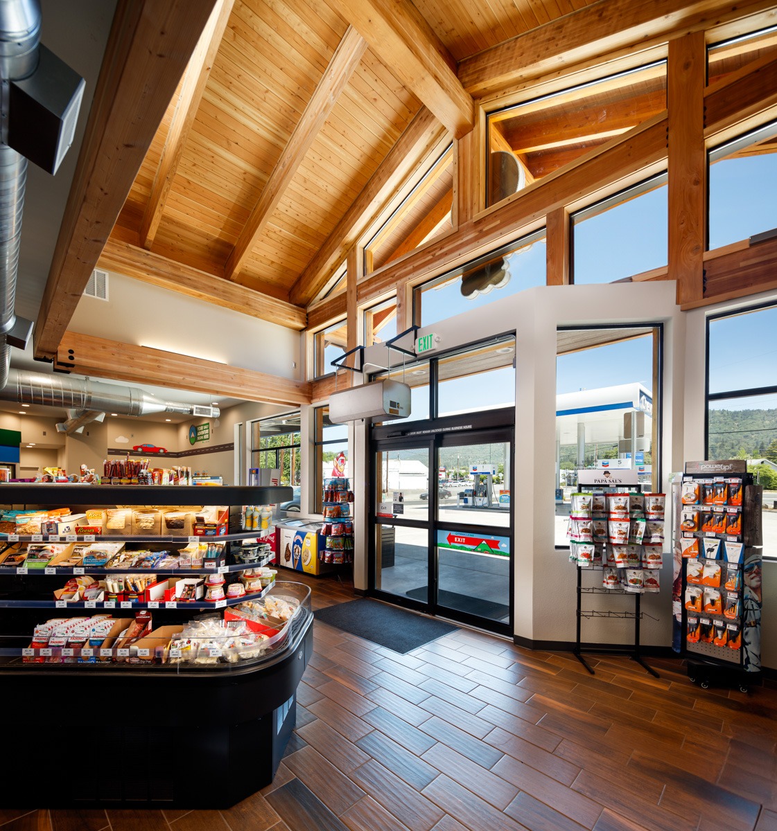 Bright, modern convenience store interior with wooden beams, featuring snacks and drinks displays, and a view of a gas station outside.